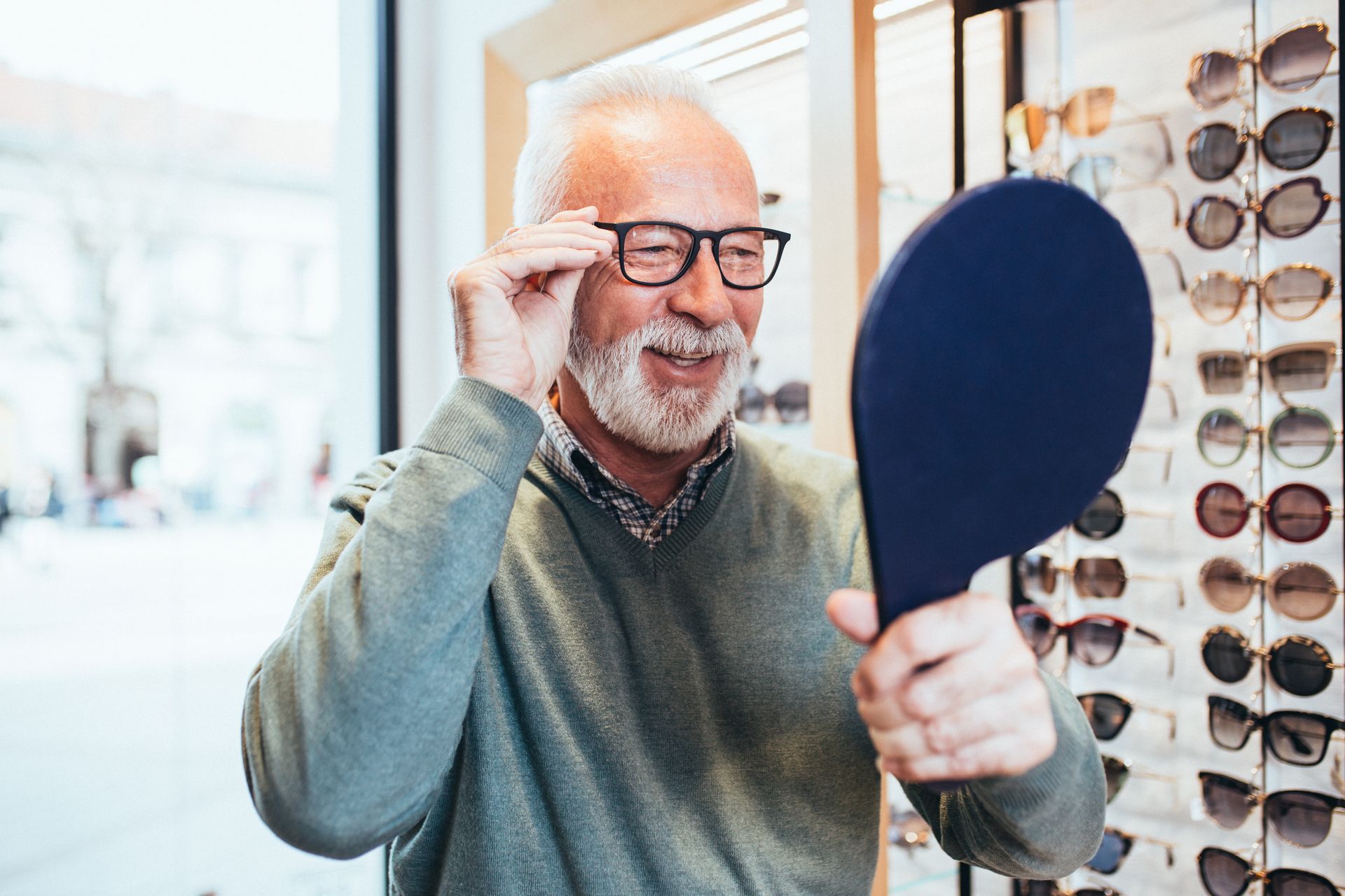 Man adjusting eyeglasses while looking into a hand mirror beside a display of eyeglass frames