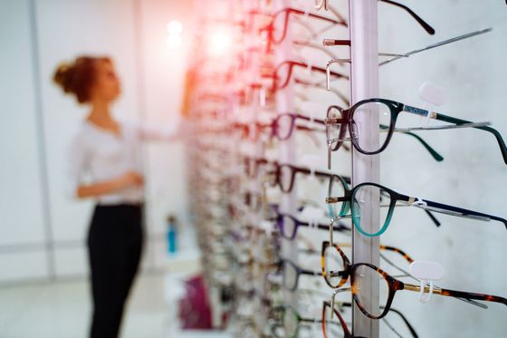 Eyeglass frames displayed on a wall, with a blurred person browsing in the background. Eyeglass frames displayed on a wall, with a blurred person browsing in the background.