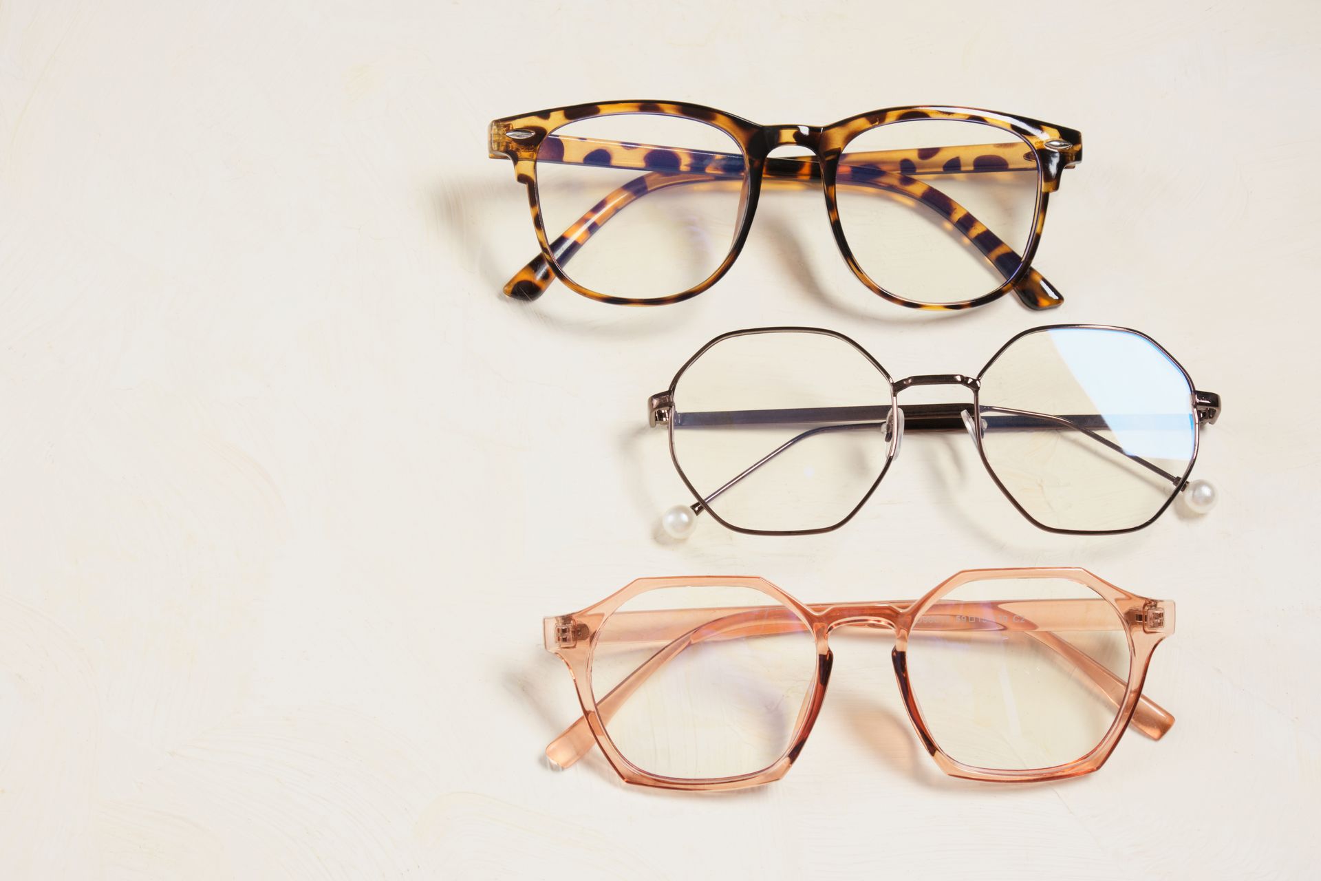 Three pairs of eyeglasses on a light background: tortoiseshell, silver, and pink frames.