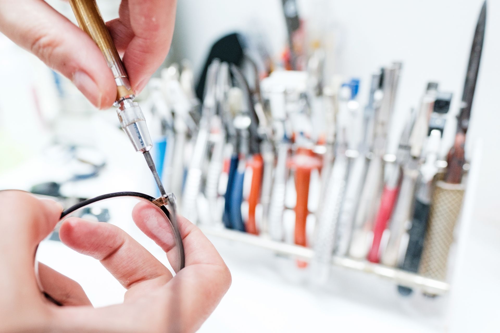 Hands holding black eyeglass frames over a white workbench with eyeglass repair tools