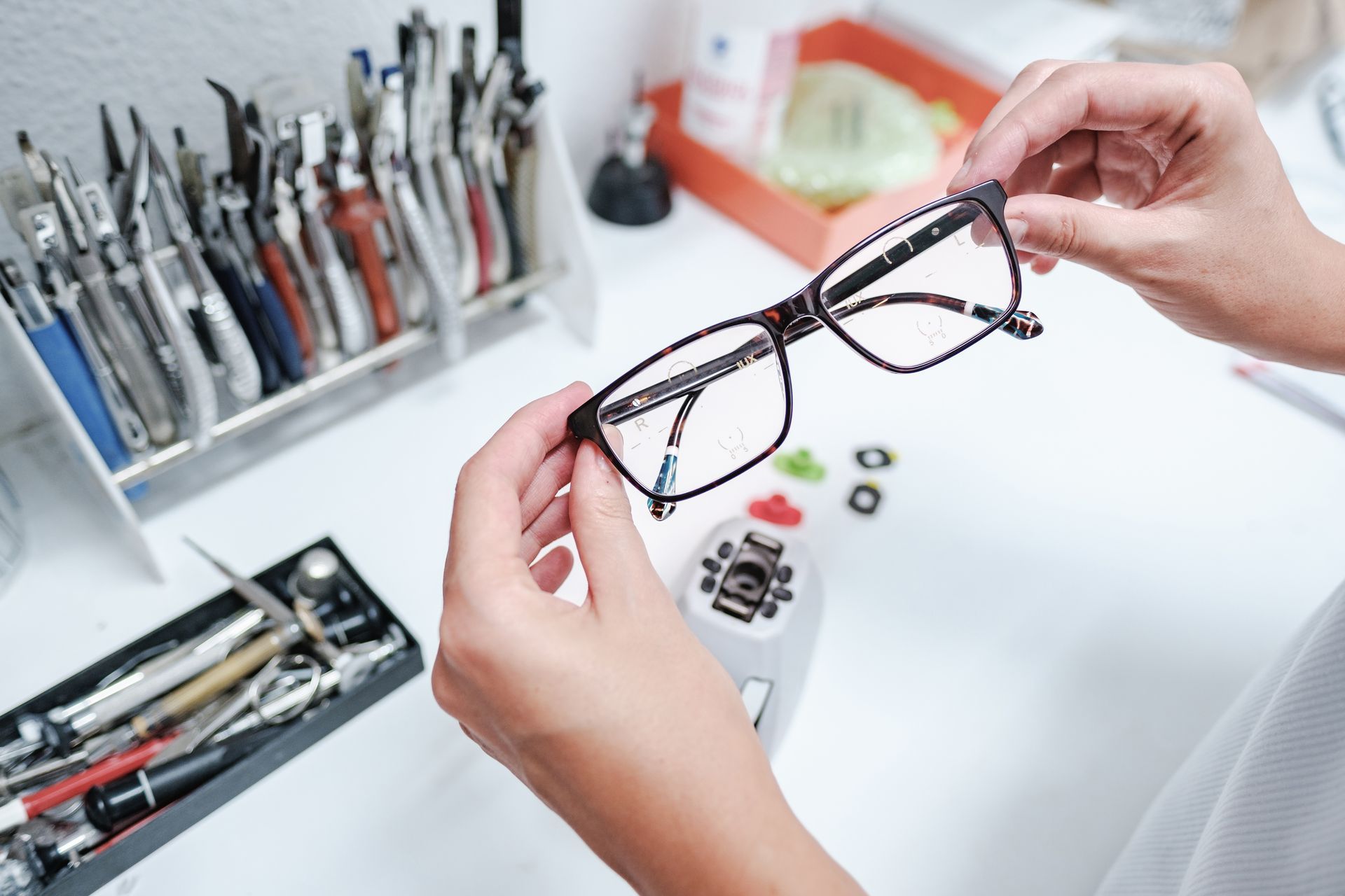Hands holding black eyeglass frames over a white workbench with eyeglass repair tools