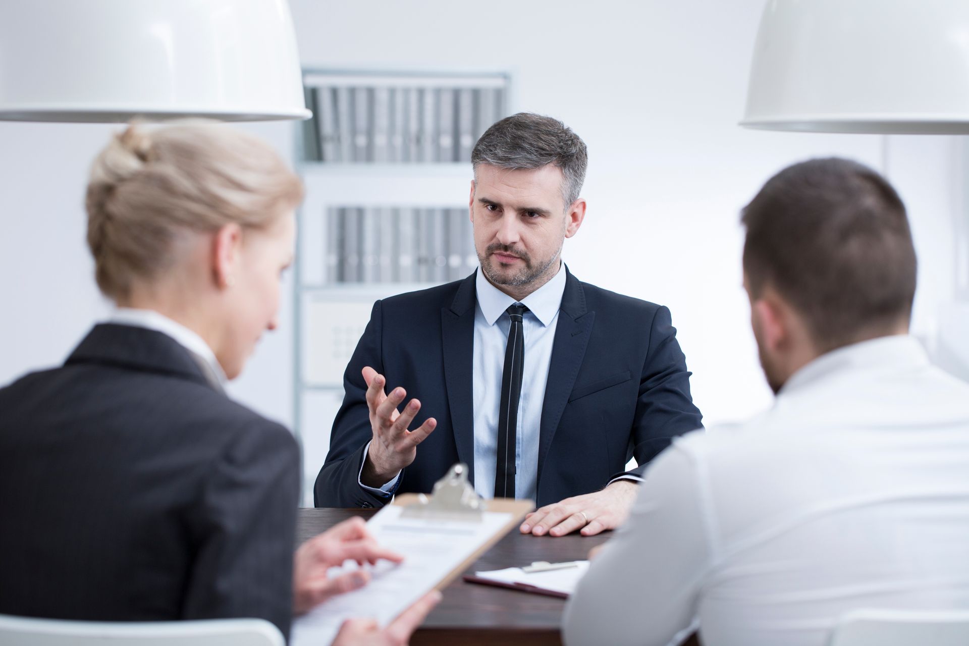 A man in a suit gestures, speaking to two people at a table; they appear to be in an office setting.
