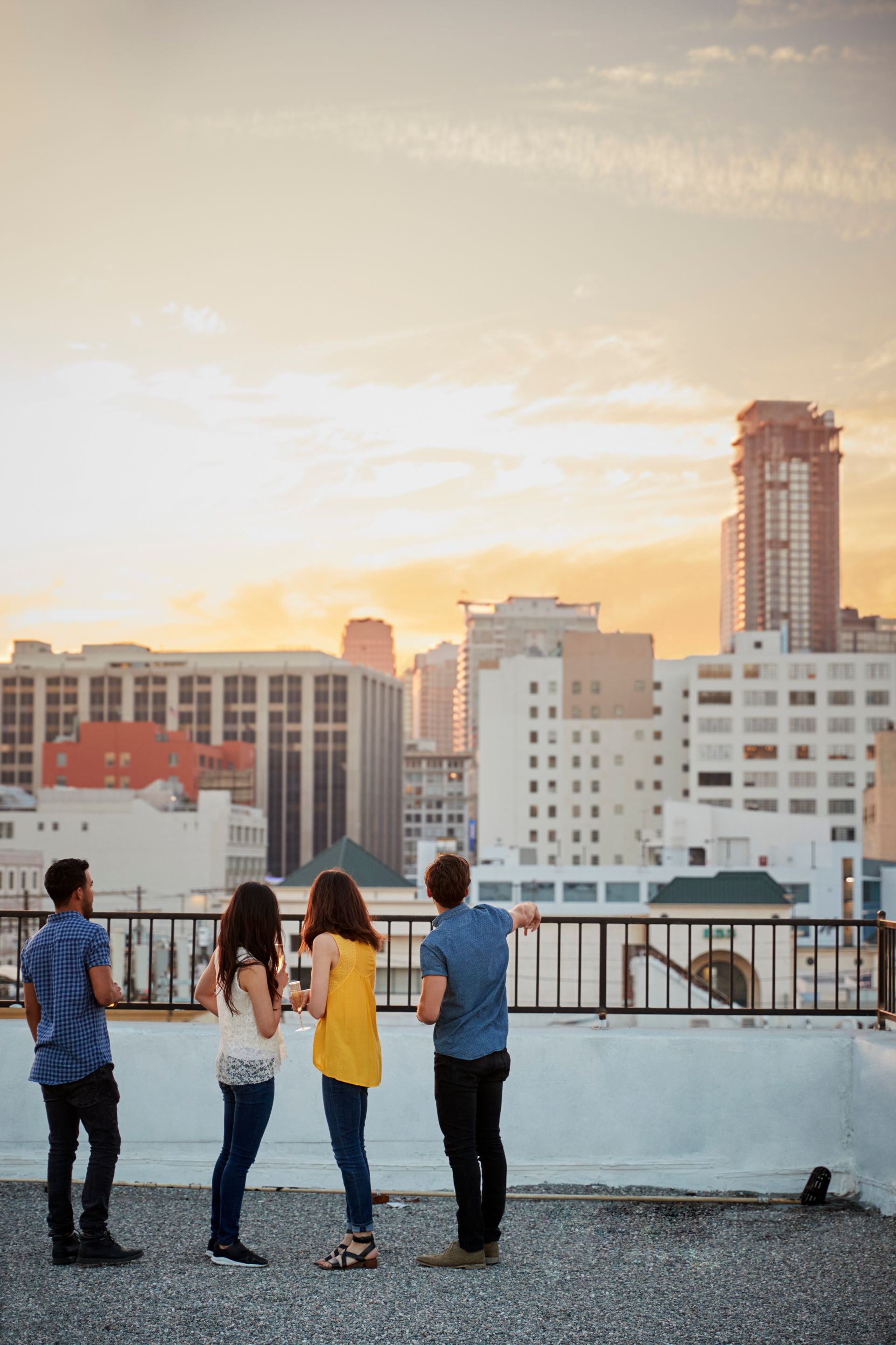 Four people on a rooftop overlooking city buildings at sunset. One points towards the horizon.