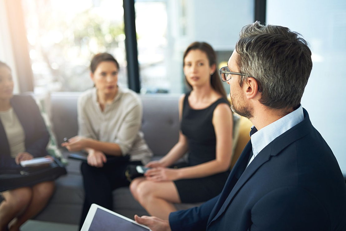Business meeting with four people sitting on a couch. The man in focus is holding a tablet.