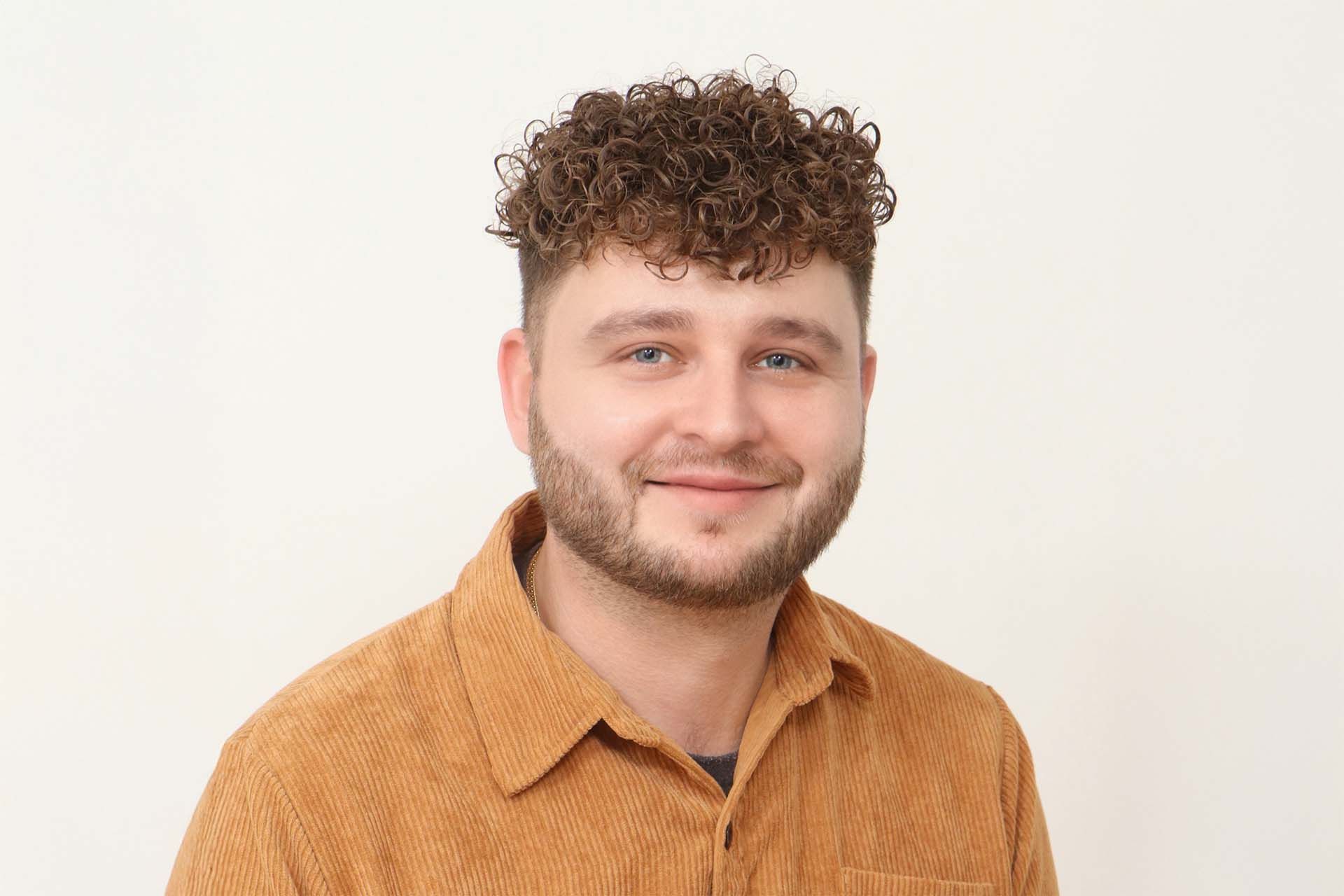 Ein Mann mit lockigem Haar und Bart trägt ein braunes Hemd.