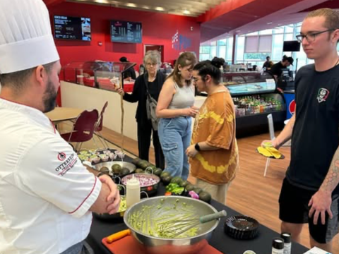 A chef demonstrating a recipe to students at a food service station in a dining hall.
