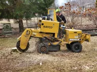 A person operating a yellow Rayco stump grinder on grass, creating wood chips.