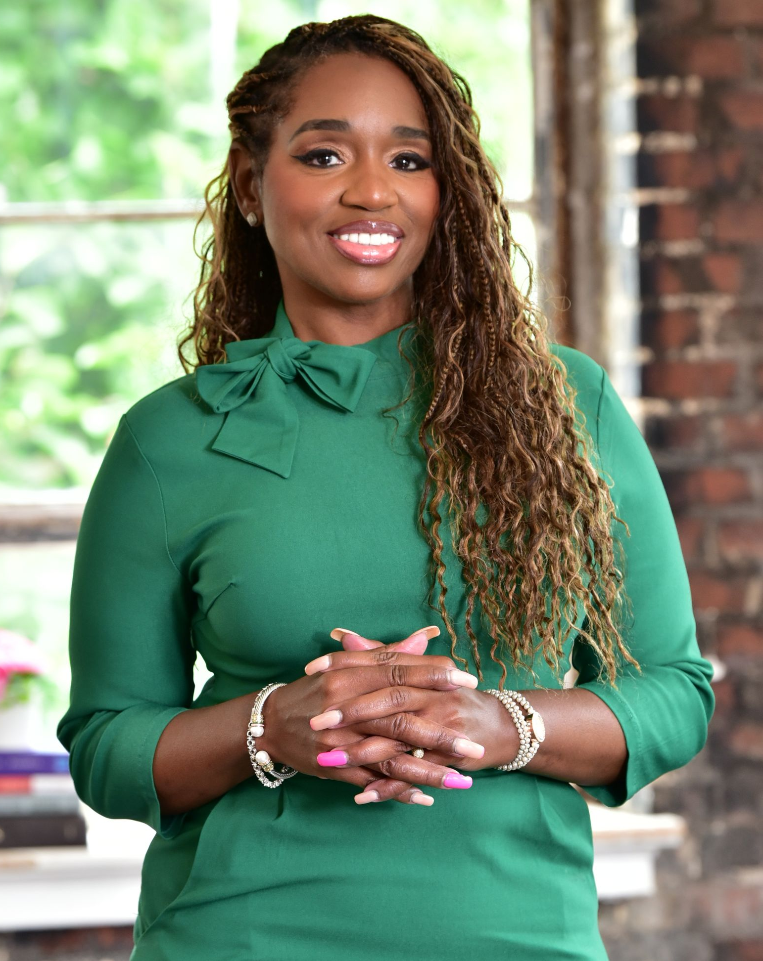 Woman in green dress, hands clasped, smiling. Background includes a window and brick wall.
