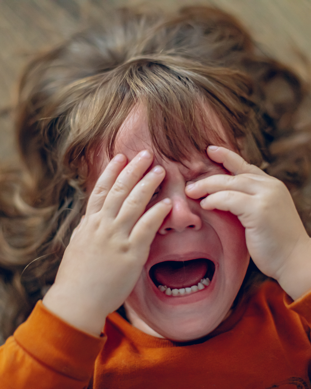 Child in an orange shirt crying and covering their eyes with both hands.