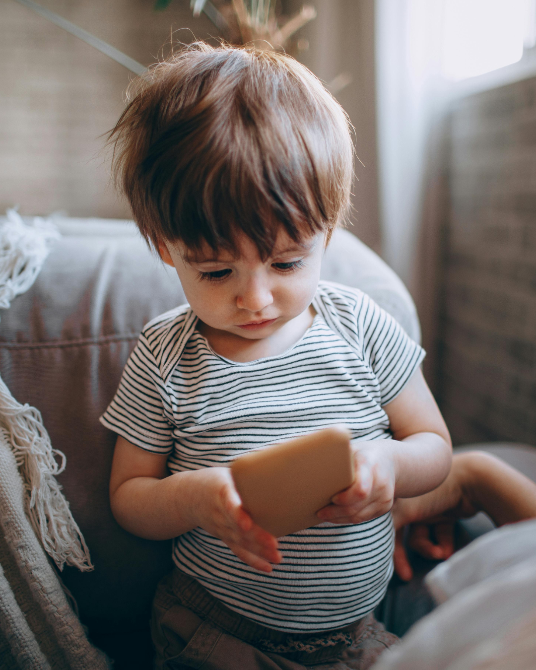 Small child sitting on a couch, looking down at a smartphone in a cozy indoor setting