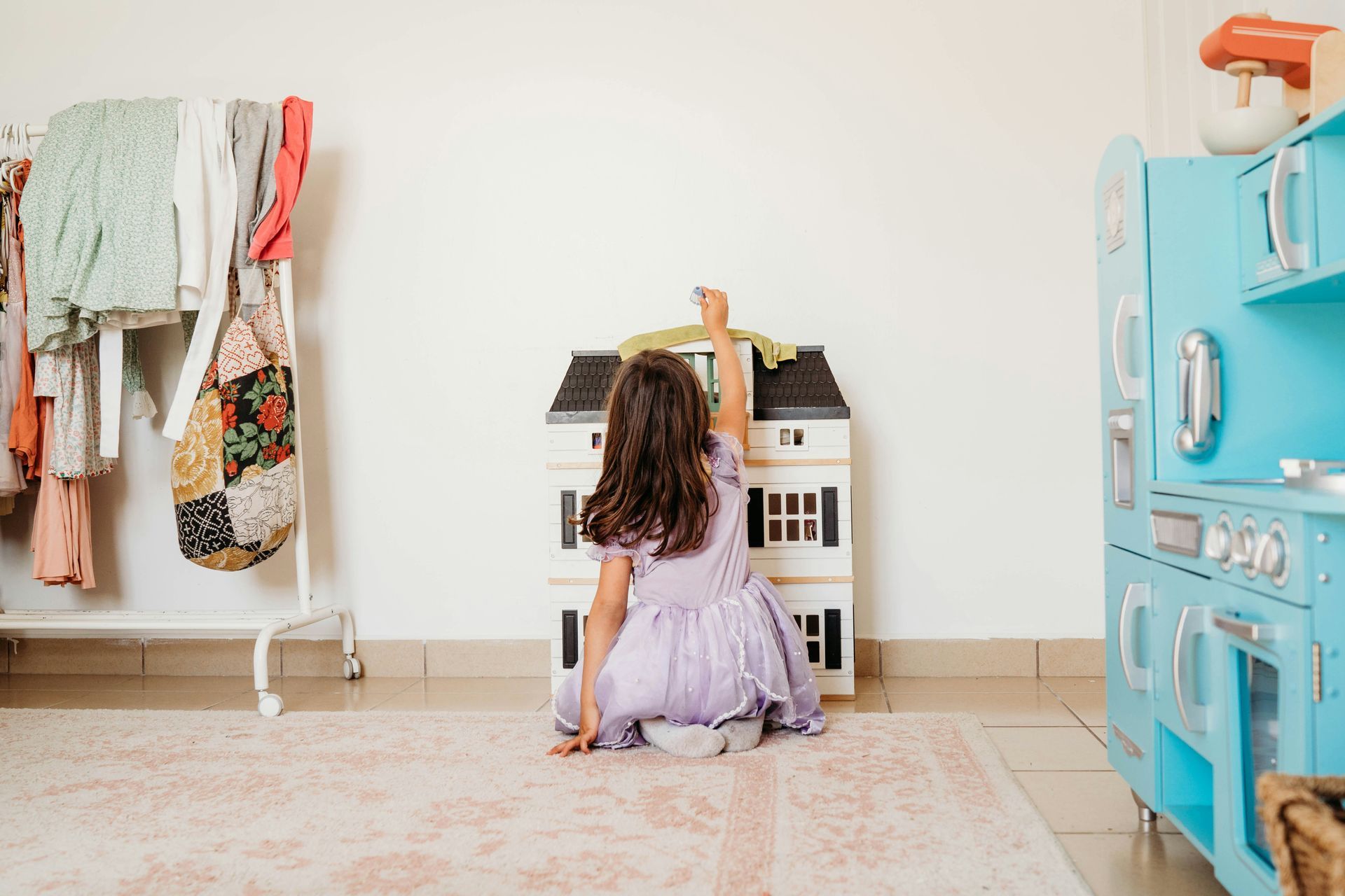 Child kneeling at a toy house in a bright playroom, with clothes on a rack and a blue toy kitchen nearby.