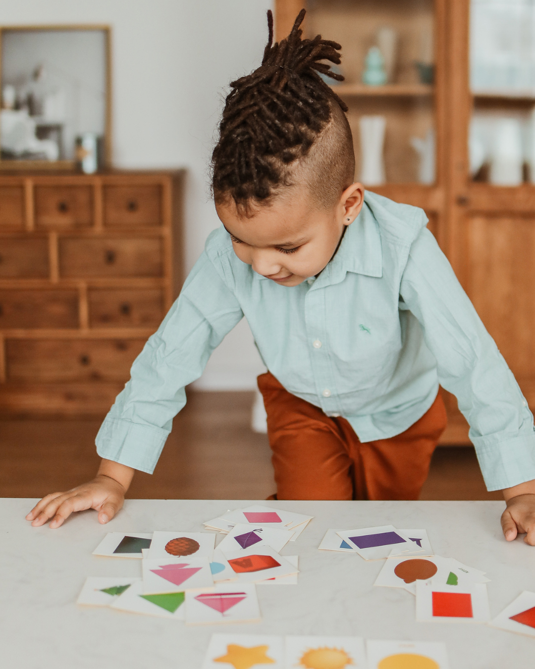 Child leaning over a table, arranging colorful paper shapes on a worksheet in a bright room