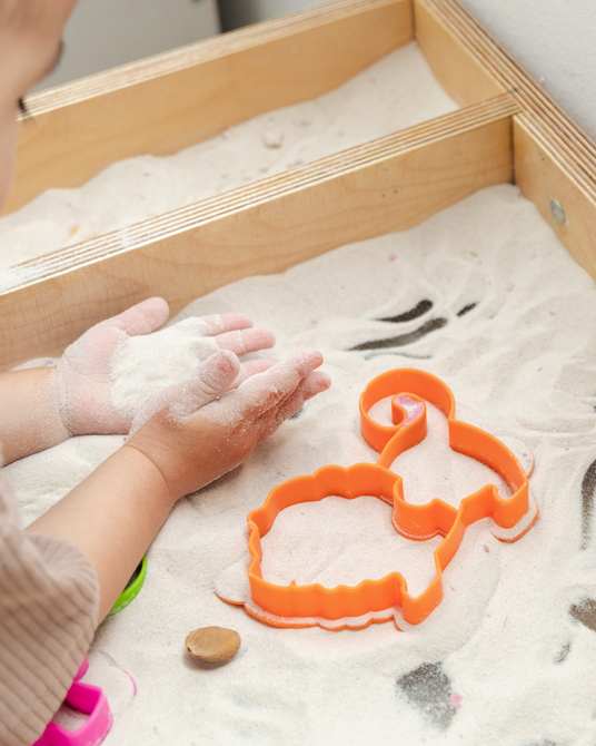 Hands in a sensory sand tray with orange cookie cutters and a small stone