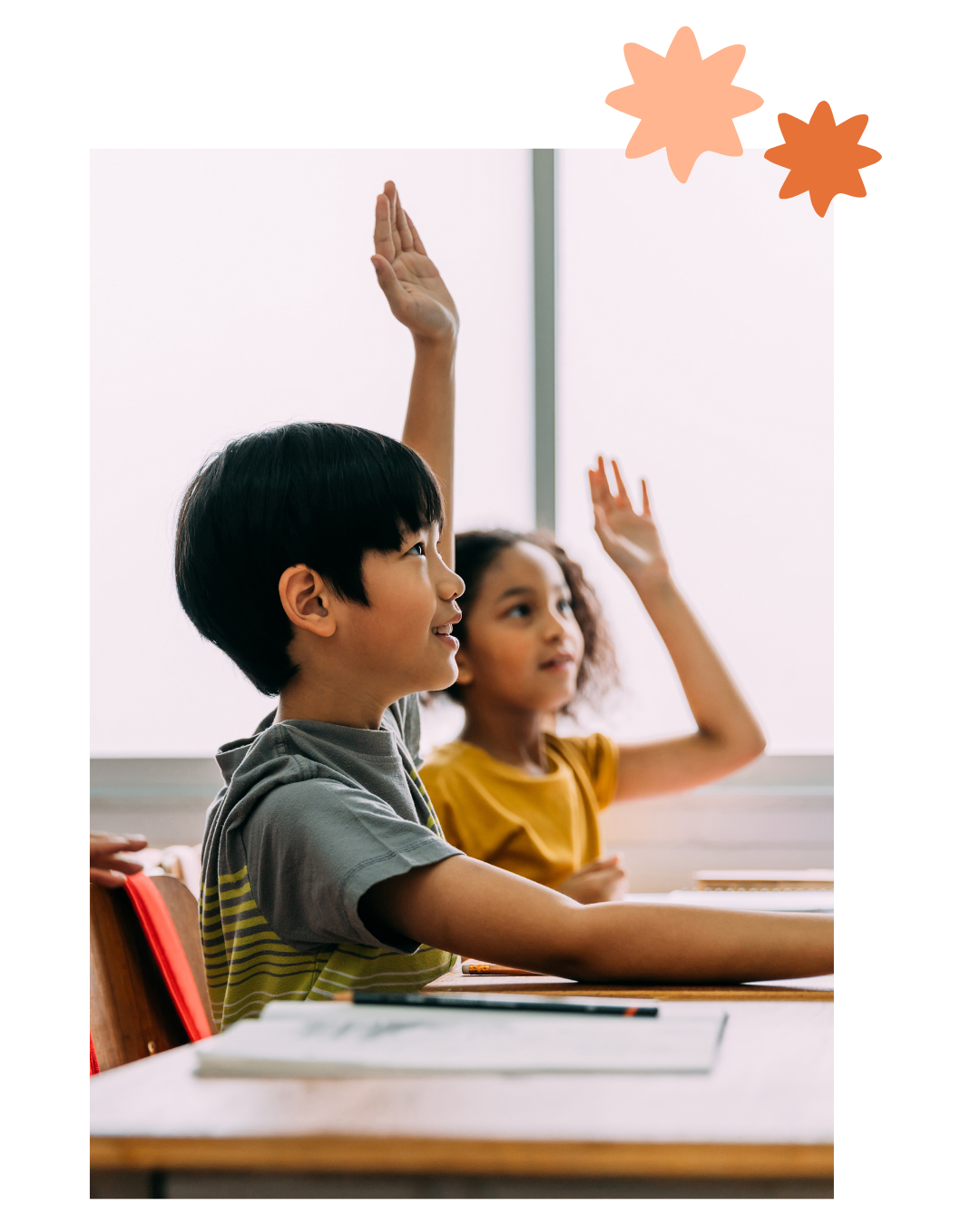 Two students raising hands in a classroom, seated at desks with papers and bright windows behind them.