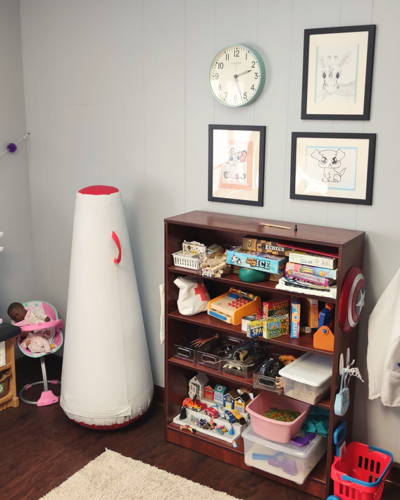 Playroom corner with toy shelf, wall clock, framed art, and white floor lamp near a doll stroller.