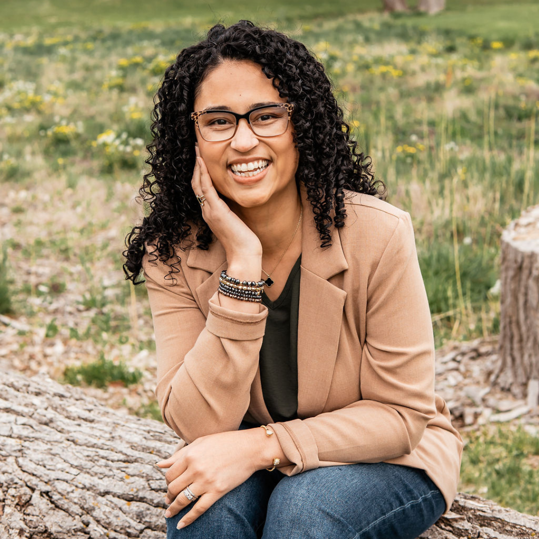 Smiling person with curly hair sitting on a log outdoors, wearing glasses, tan jacket, and jeans.