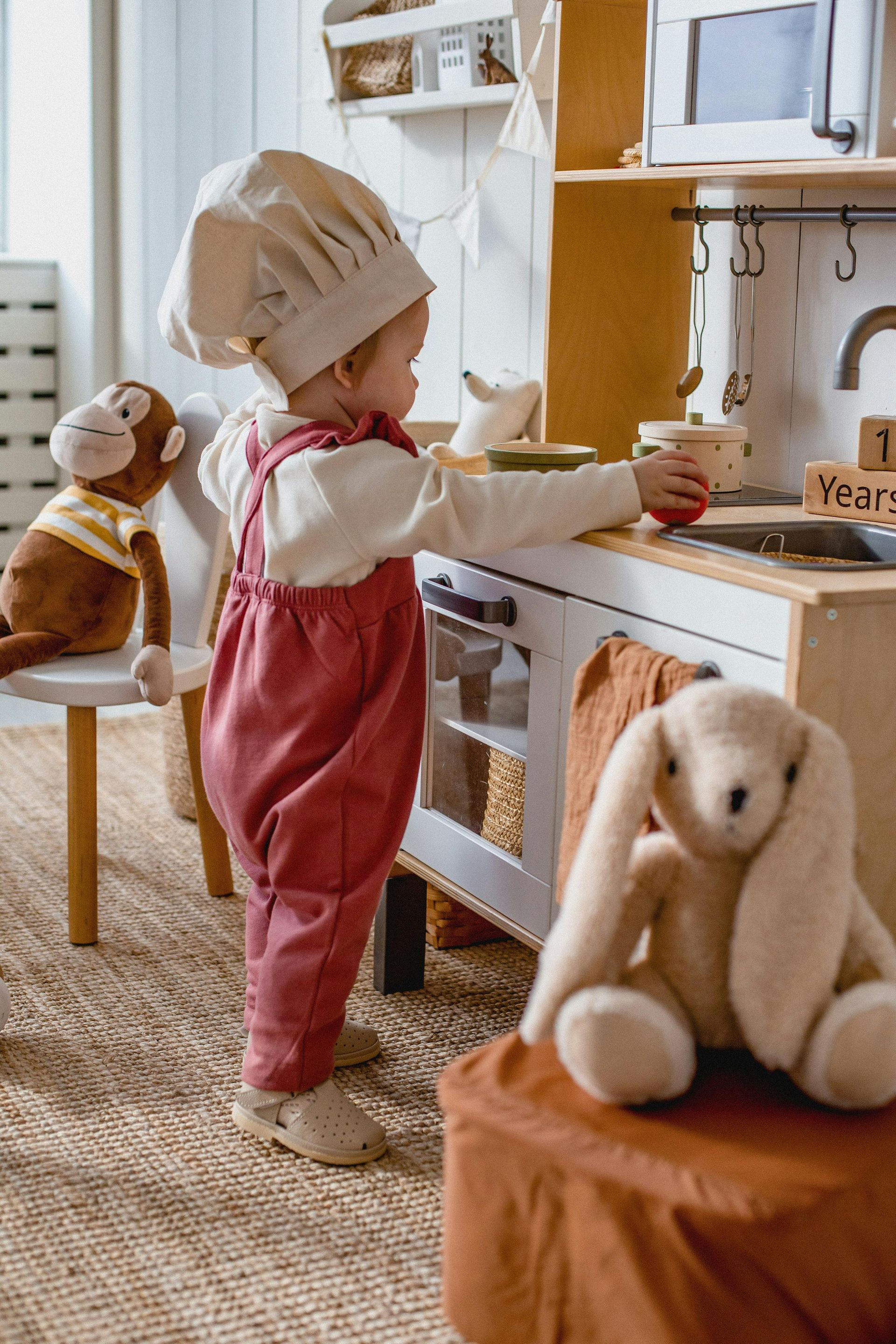 Child in pink overalls and chef hat playing in a toy kitchen with stuffed animals.