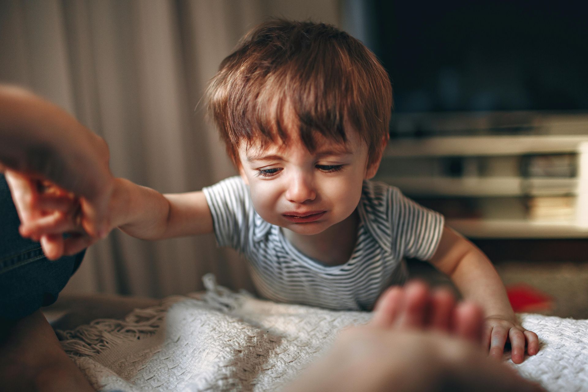 Child reaching for an adult’s hand on a bed, with a soft, warm indoor background