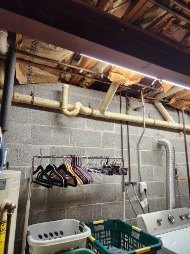 A basement laundry area with exposed overhead pipes and insulation, a clothing rack, laundry baskets, and a dryer.