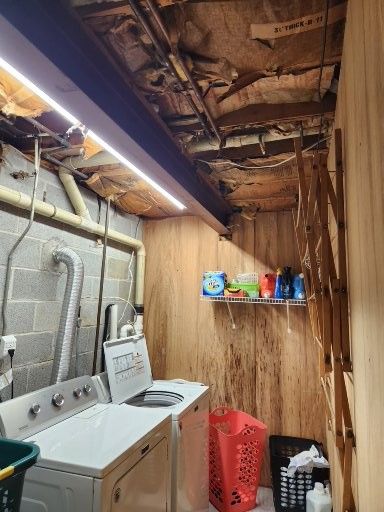 A basement laundry area with a white washer and dryer, a wire shelf with supplies, a red laundry basket, and plywood walls.