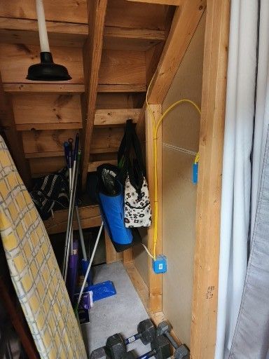 A cramped storage nook under wooden stairs containing cleaning supplies, a blue bag, and exposed yellow electrical wiring.