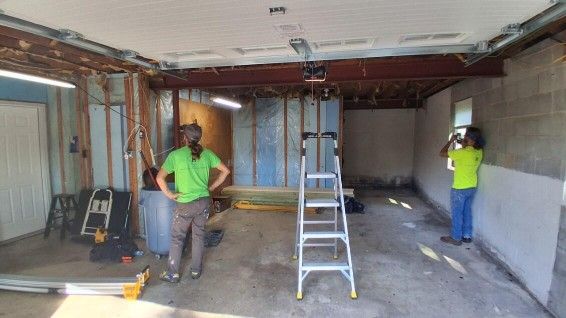 Two people in bright shirts stand in an unfinished garage with bare wall studs, a ladder, and exposed ceiling beams.