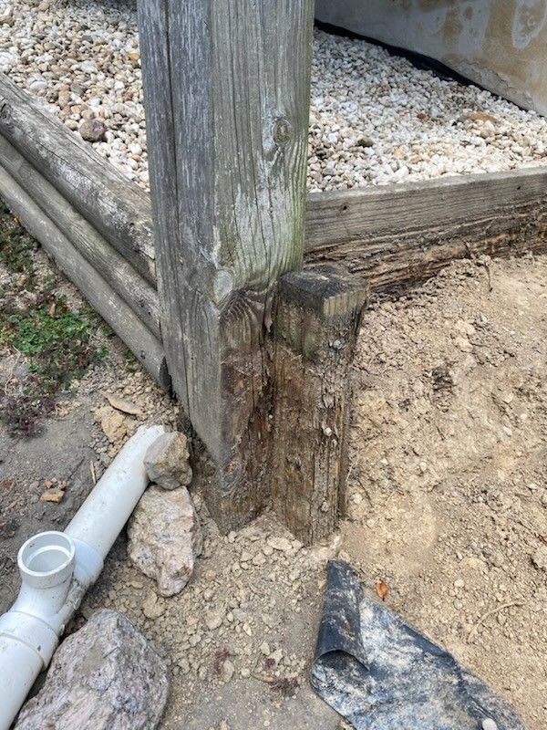 A weathered wooden support post stands next to a short, decaying stake, with loose gravel and a PVC pipe in the foreground.