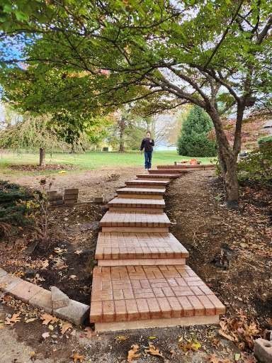 A set of new, light brown brick stairs leads up a gentle slope toward a person standing in a grassy park area.