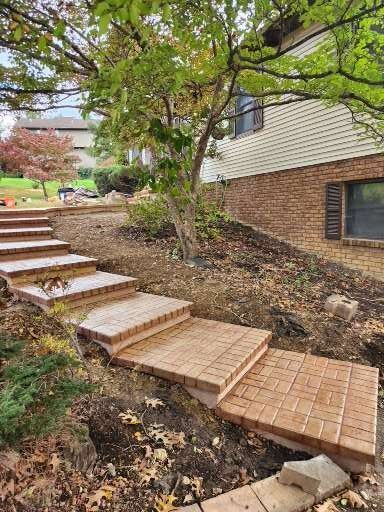 Brick steps ascend a landscaped hill alongside a house with beige siding and a brick lower level.