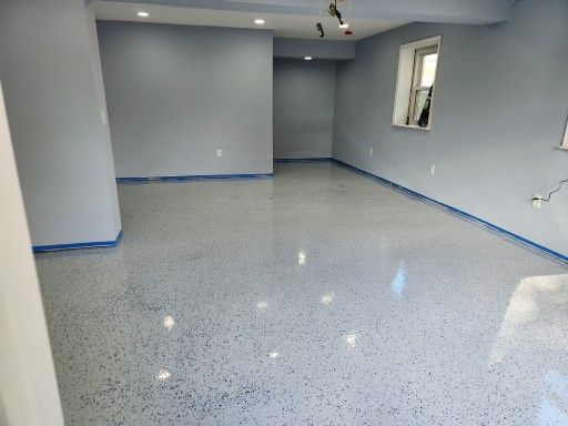 A light gray basement room with a freshly coated epoxy floor featuring blue and gray flecks, still taped for painting.
