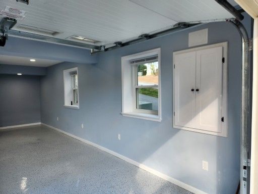 A garage interior with grey walls, a speckled floor, two windows, and a built-in white cabinet on the wall.