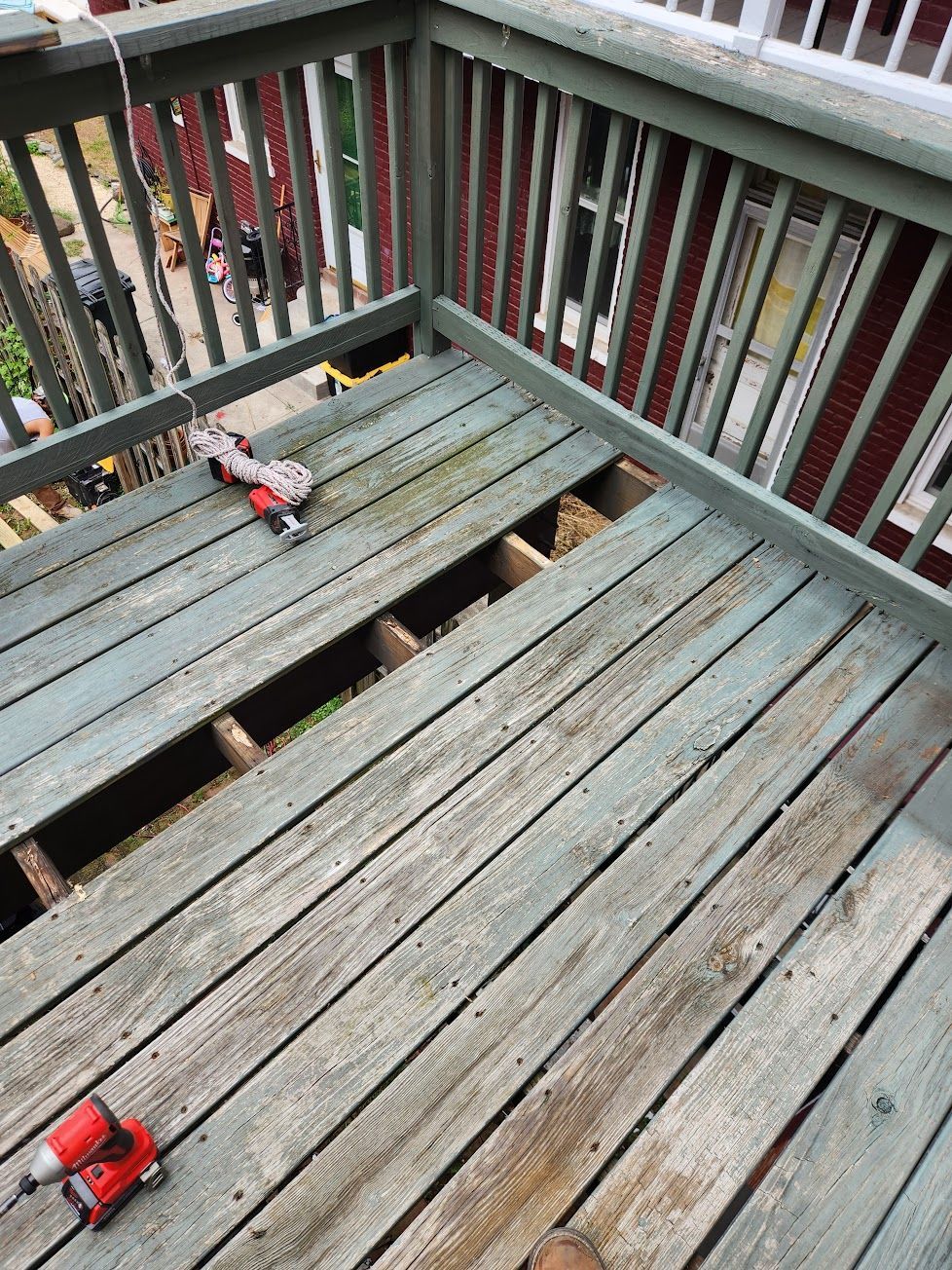 A gray wooden deck with a section of missing floorboards, showing the joists underneath, with a red power drill nearby.