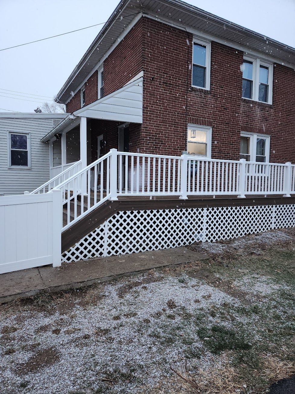 A red brick two-story house with a white porch, white lattice skirting, and white vinyl fencing in a snowy setting.