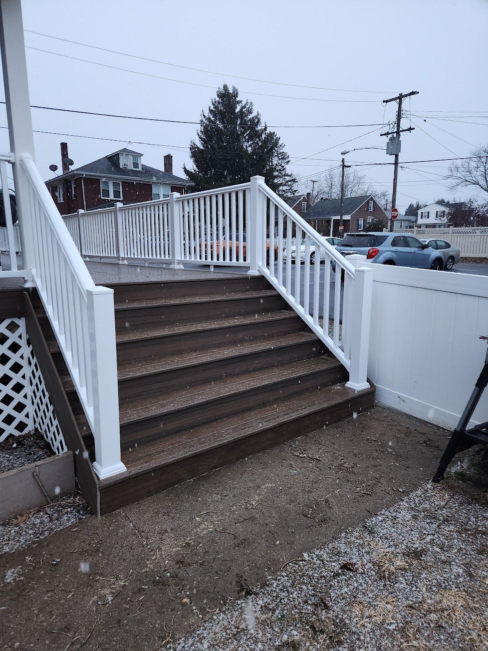 Brown wooden outdoor steps with white railings lead up to a deck, set against a background of houses and a street.