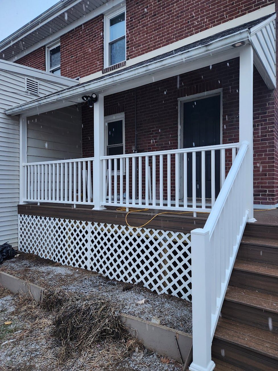 A red brick house features a raised wooden porch with white railings, stairs, and lattice skirting under a metal roof.