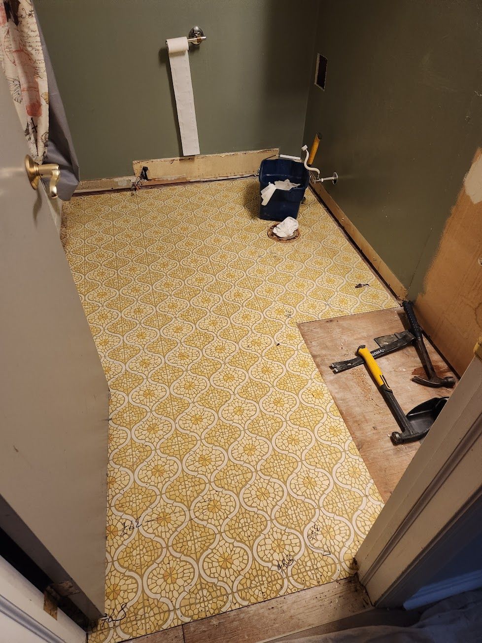 A bathroom floor under renovation with patterned yellow vinyl flooring, a toilet flange, and tools on a bare subfloor.