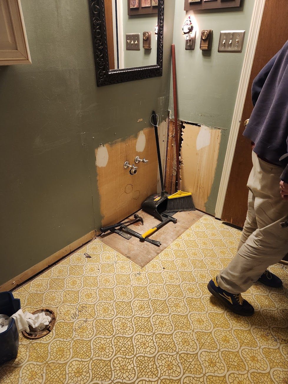 A person stands in a bathroom with removed vanity cabinetry, exposing wall studs and pipes above yellow patterned flooring.