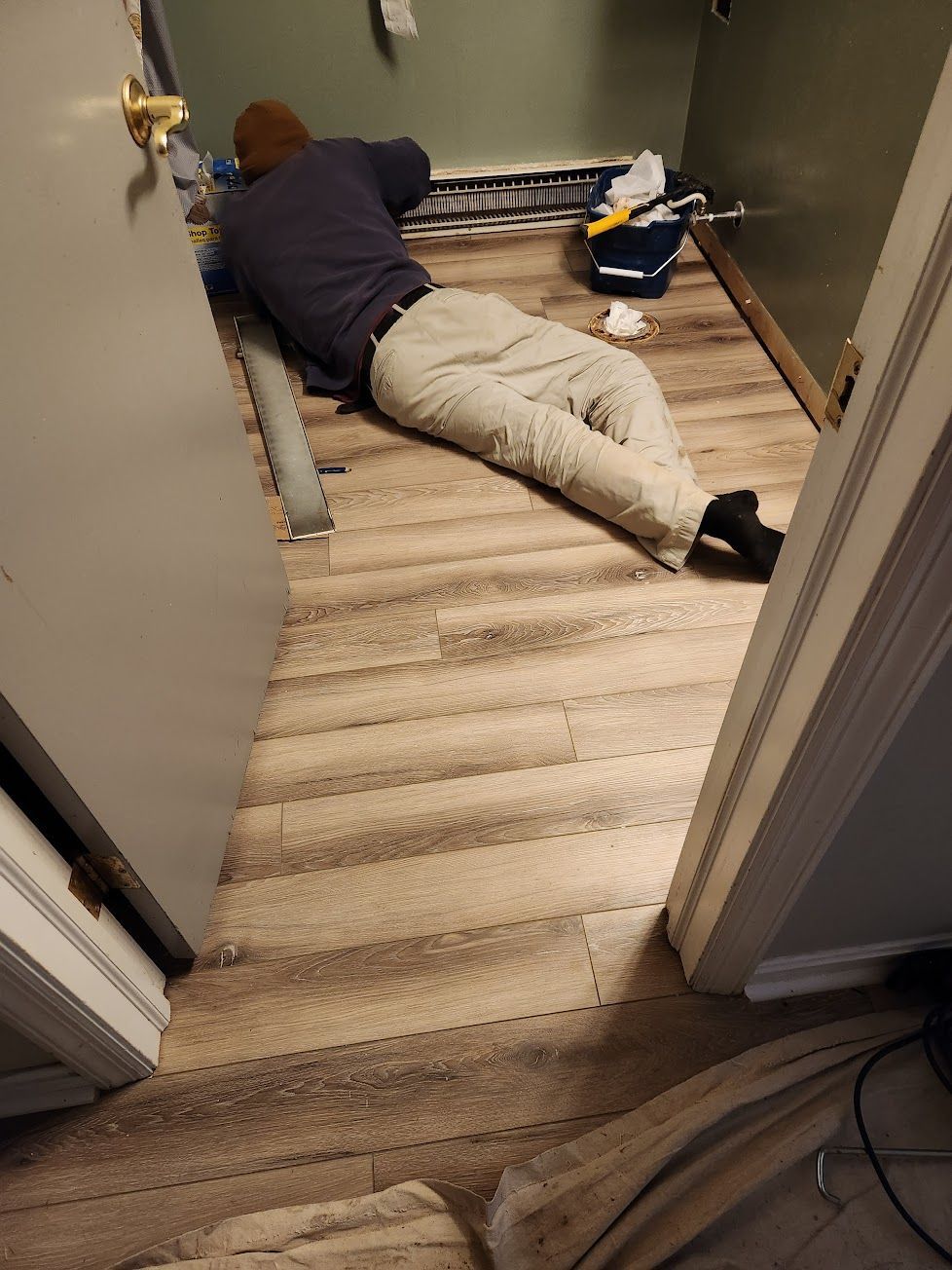 A person in a dark sweater and light pants lying on the floor, installing vinyl plank flooring in a room.