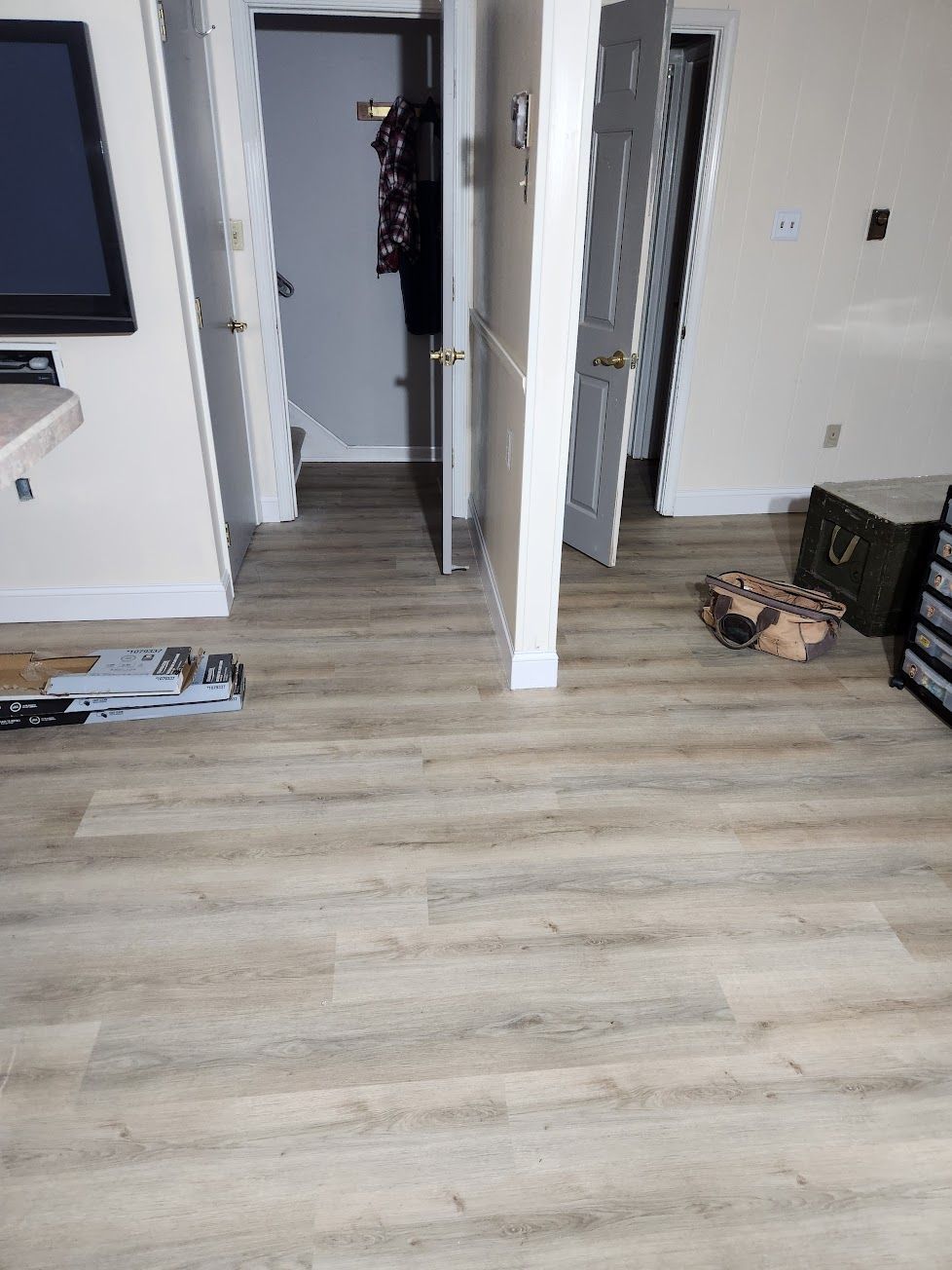 A bright room with light wood-look flooring features two open doorways, a wall-mounted TV, and a box on the floor.