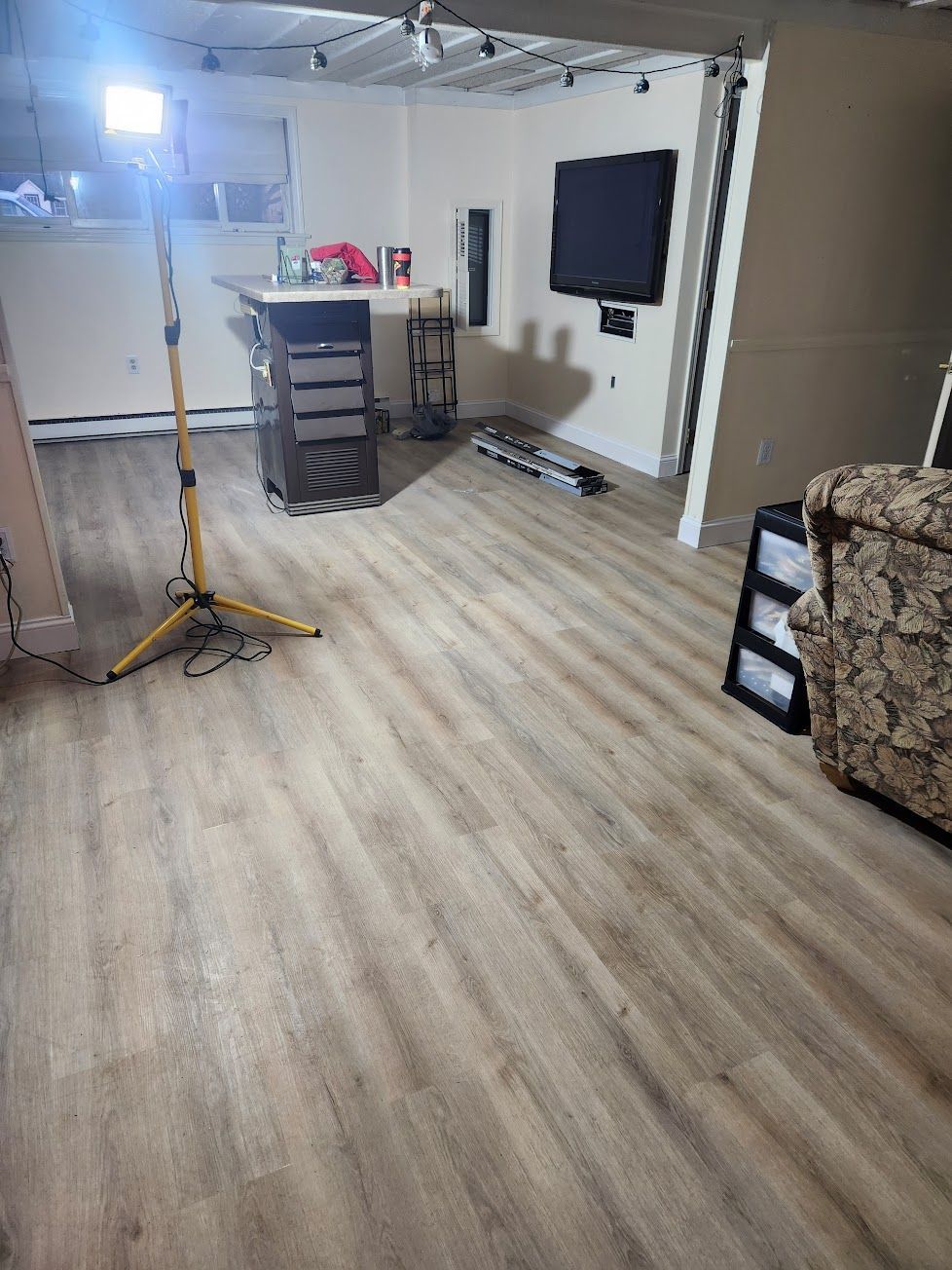 A brightly lit room with light wood-patterned flooring, a tall work light, a small workstation, and a wall-mounted TV.