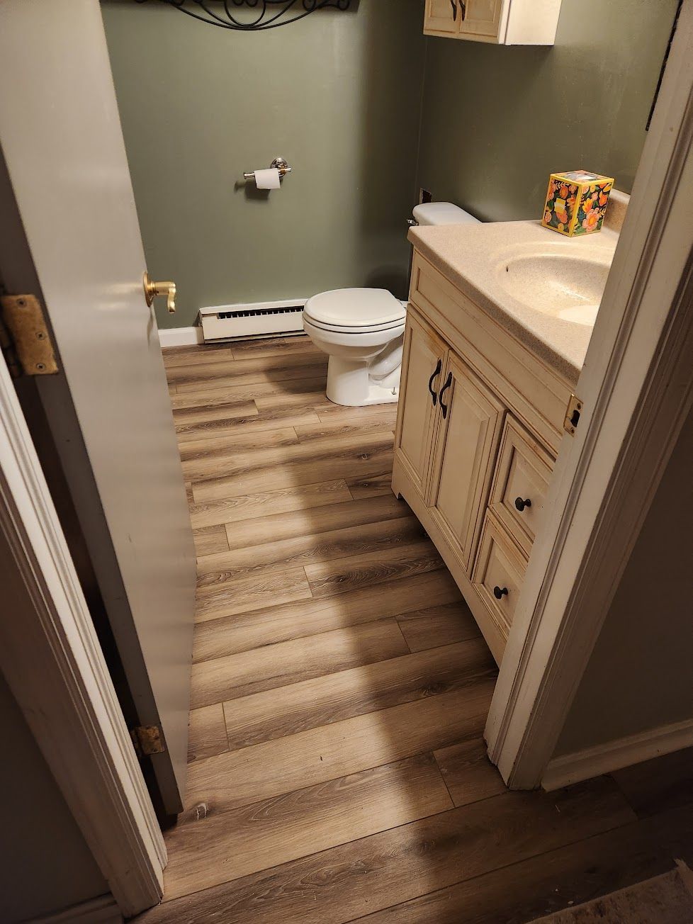 A small bathroom with green walls, wood-look flooring, a toilet, and a white vanity with a sink.
