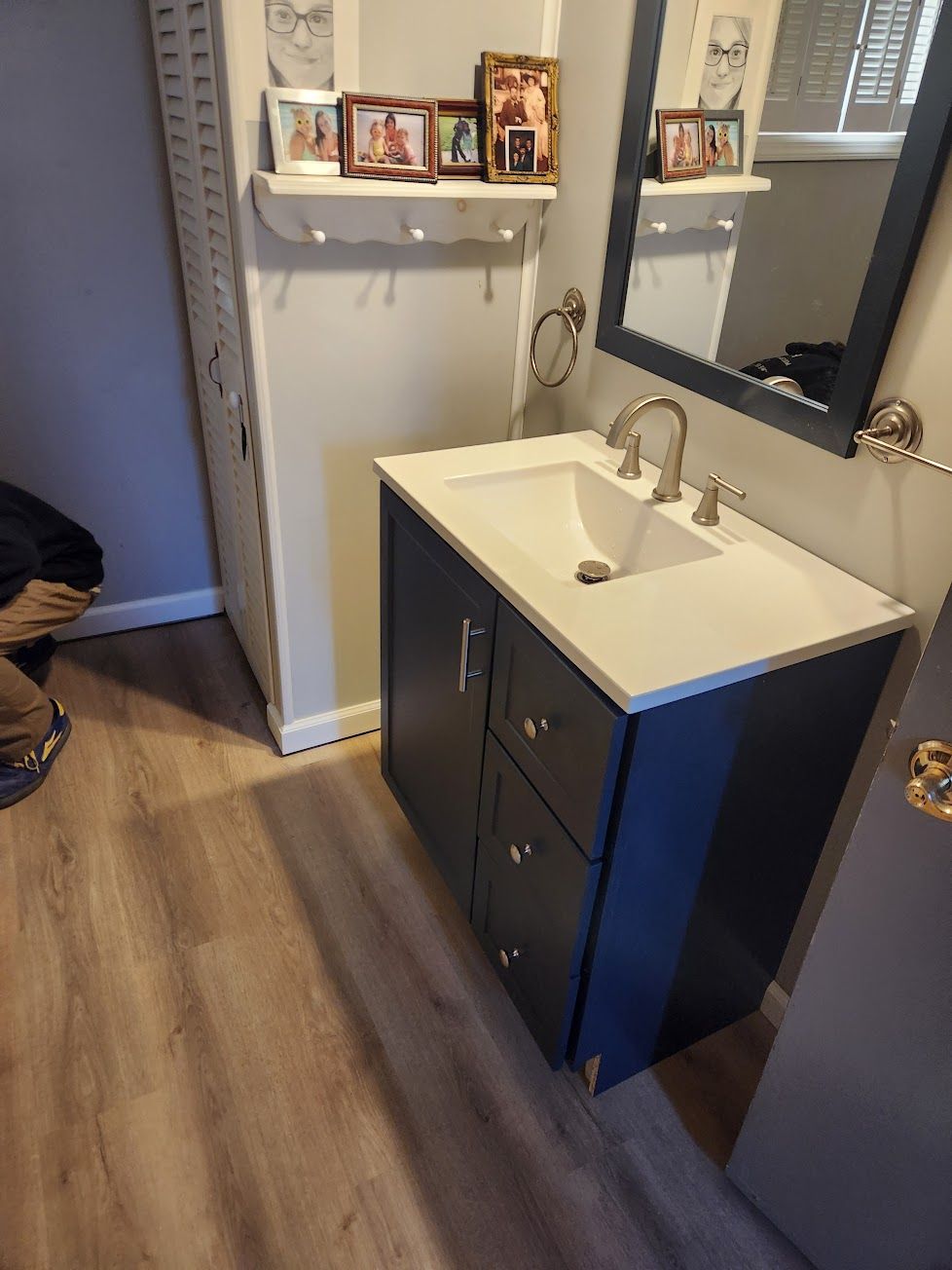 A dark blue bathroom vanity with a white countertop and sink, featuring a mirror, wall shelf with photos, and wood flooring.