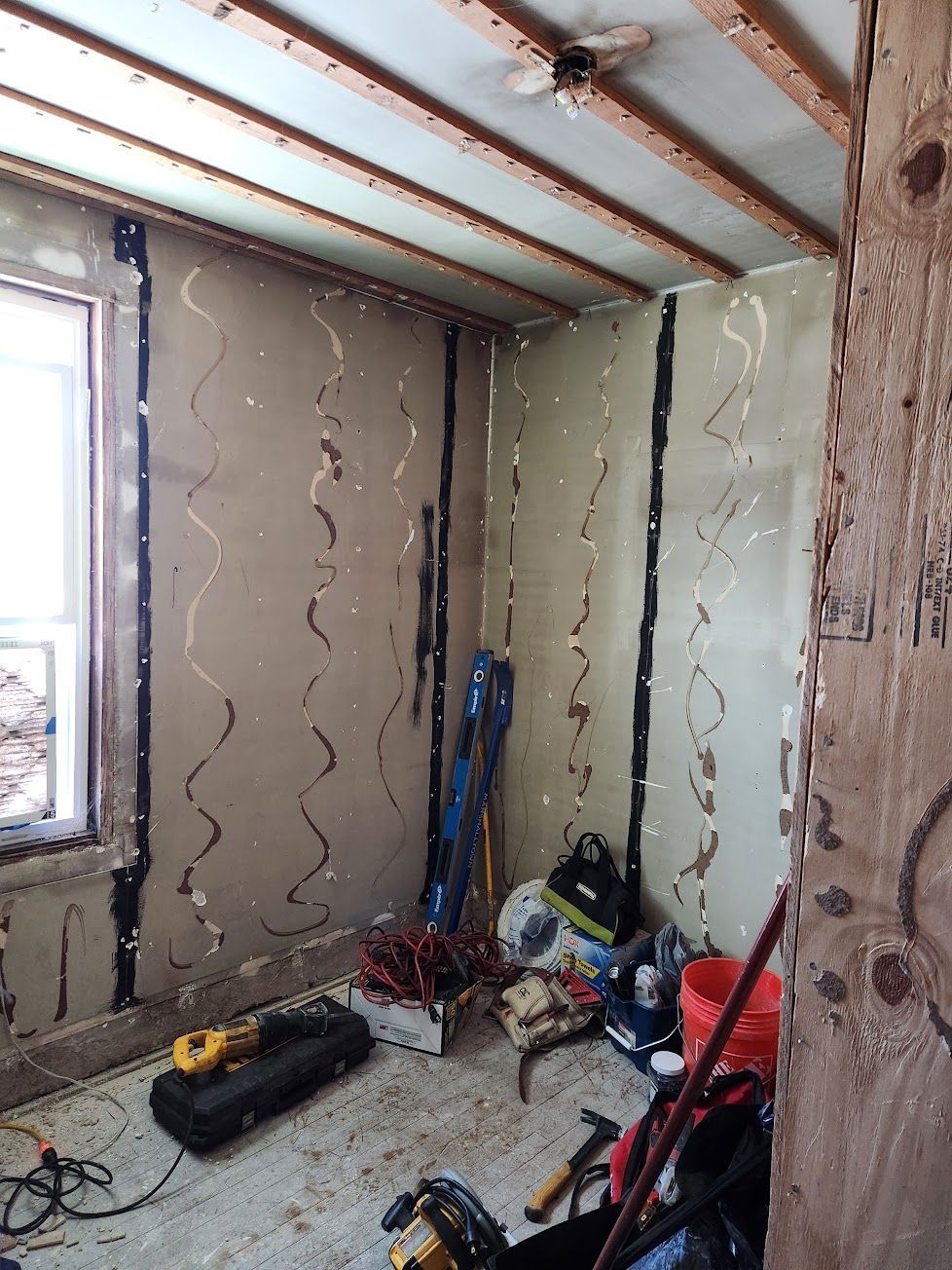 A room under renovation with bare walls showing vertical adhesive strips, exposed ceiling joists, and scattered tools.