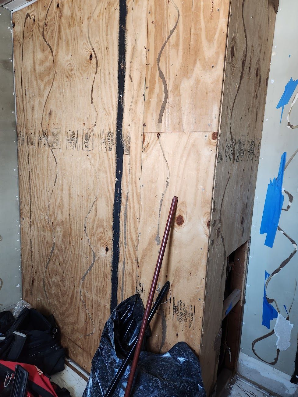 Plywood boards cover a wall section in a room undergoing renovation, with a dark seam and tools nearby.