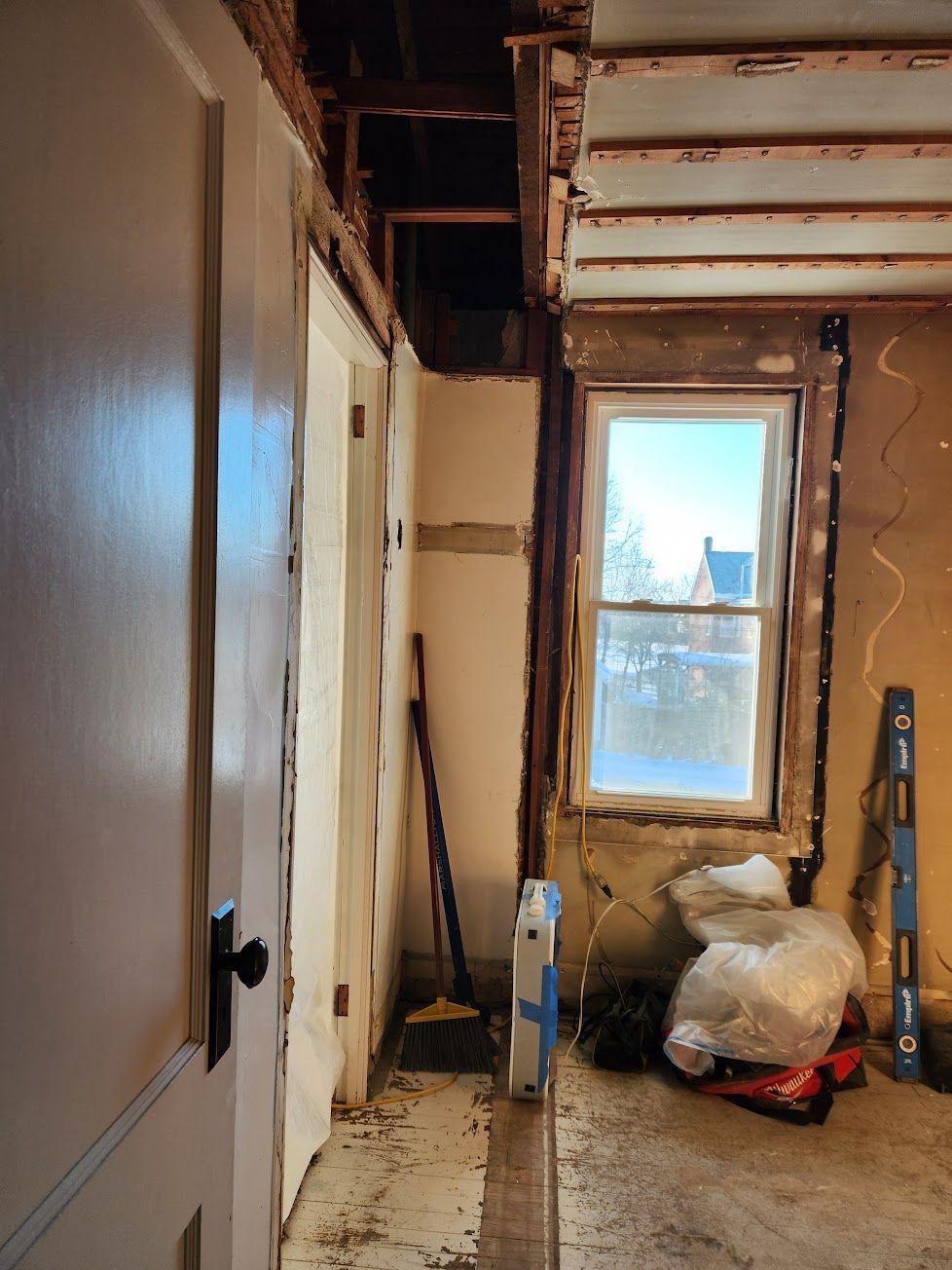 Interior view of a room under renovation with exposed ceiling joists, a window, debris, and tools on a concrete floor.