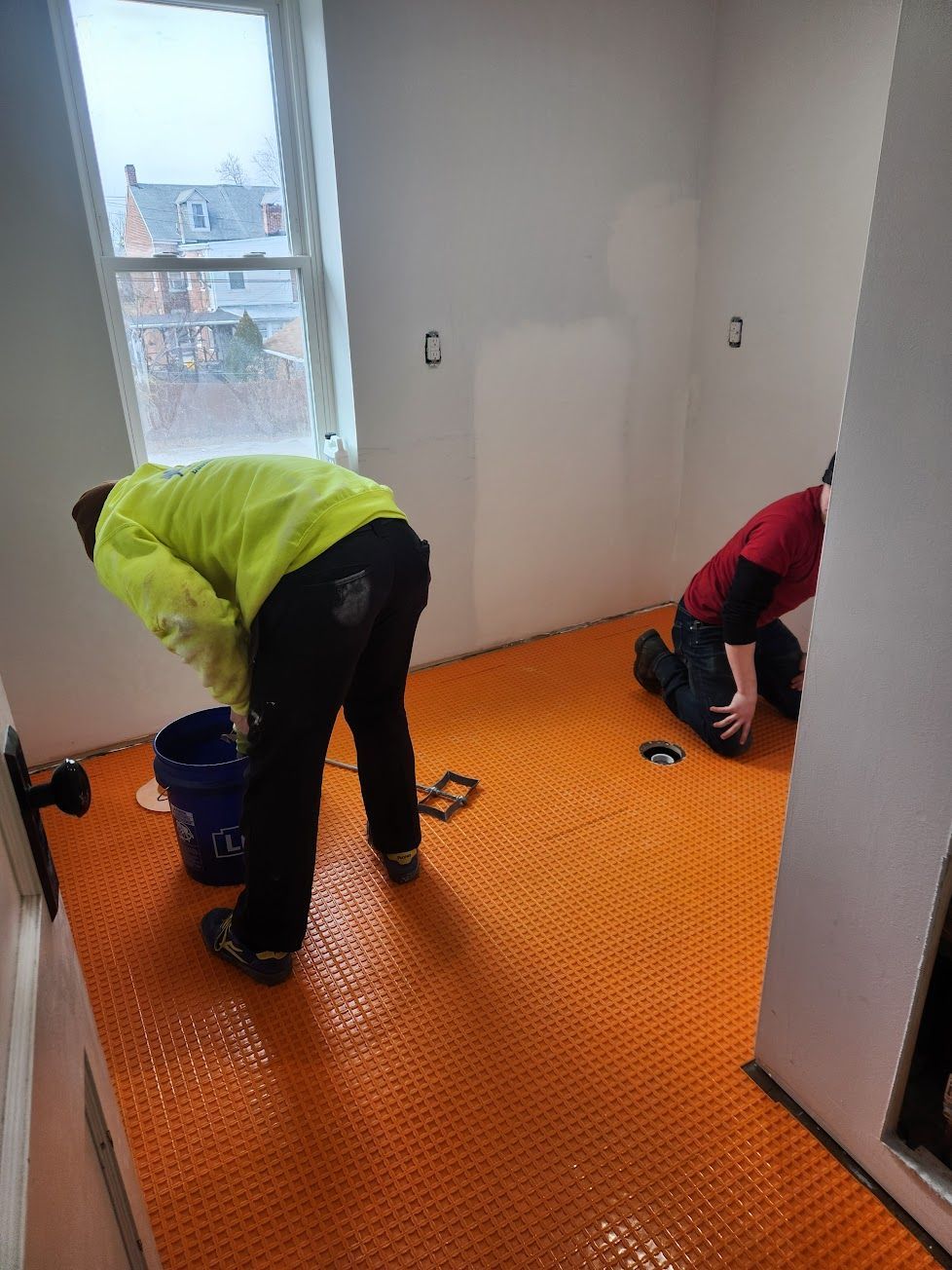 Two people install bright orange Schluter-DITRA floor underlayment in a room with a large window and bare drywall.
