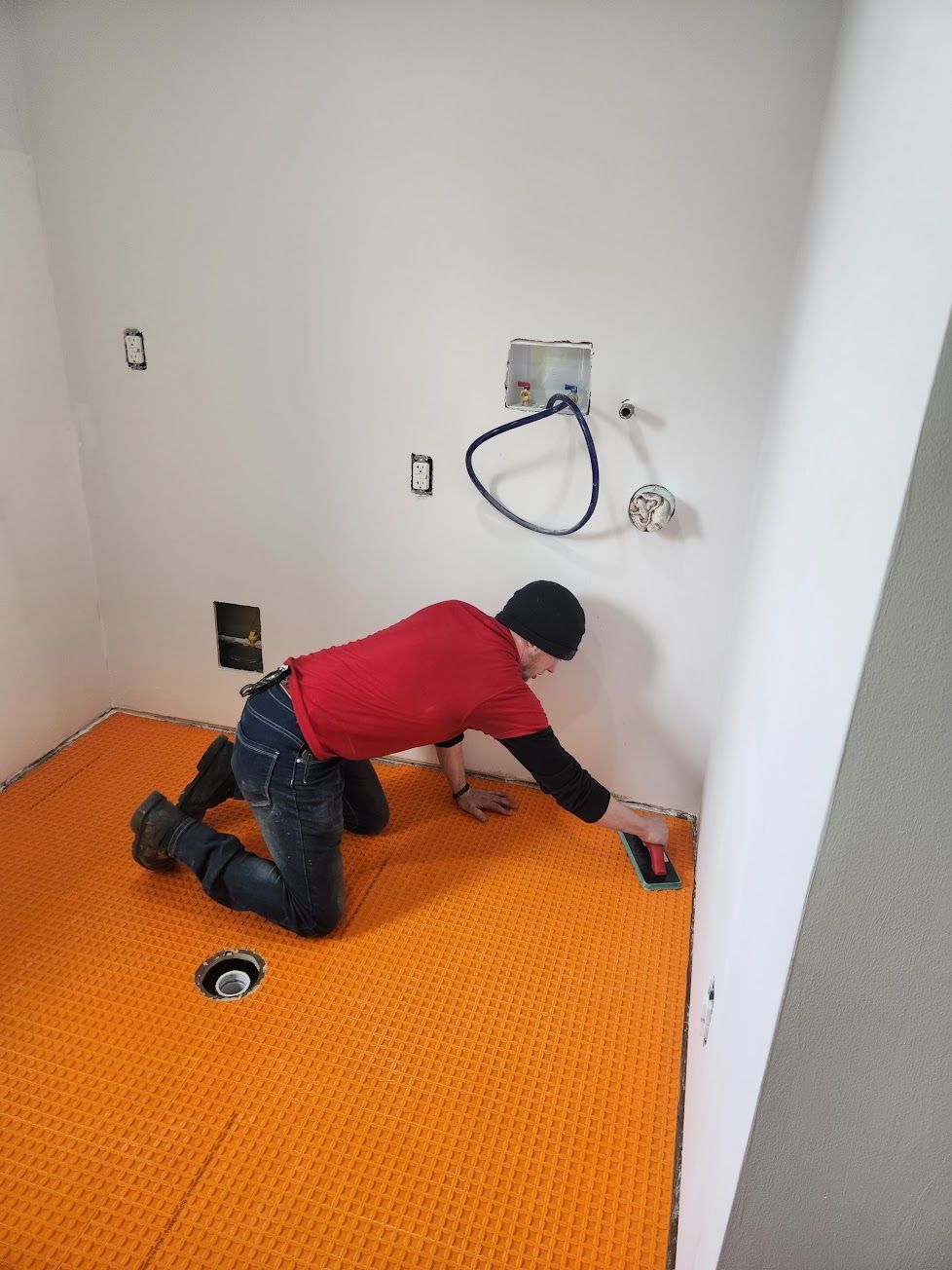 A worker in a red shirt kneels on a floor, smoothing orange uncoupling membrane onto the subfloor with a trowel.