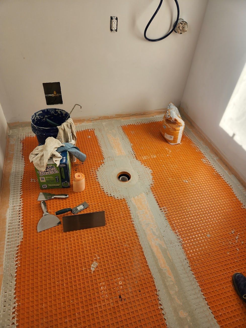 An orange Schluter-DITRA uncoupling membrane installed on a floor with a central drain, tiling tools, and supplies.