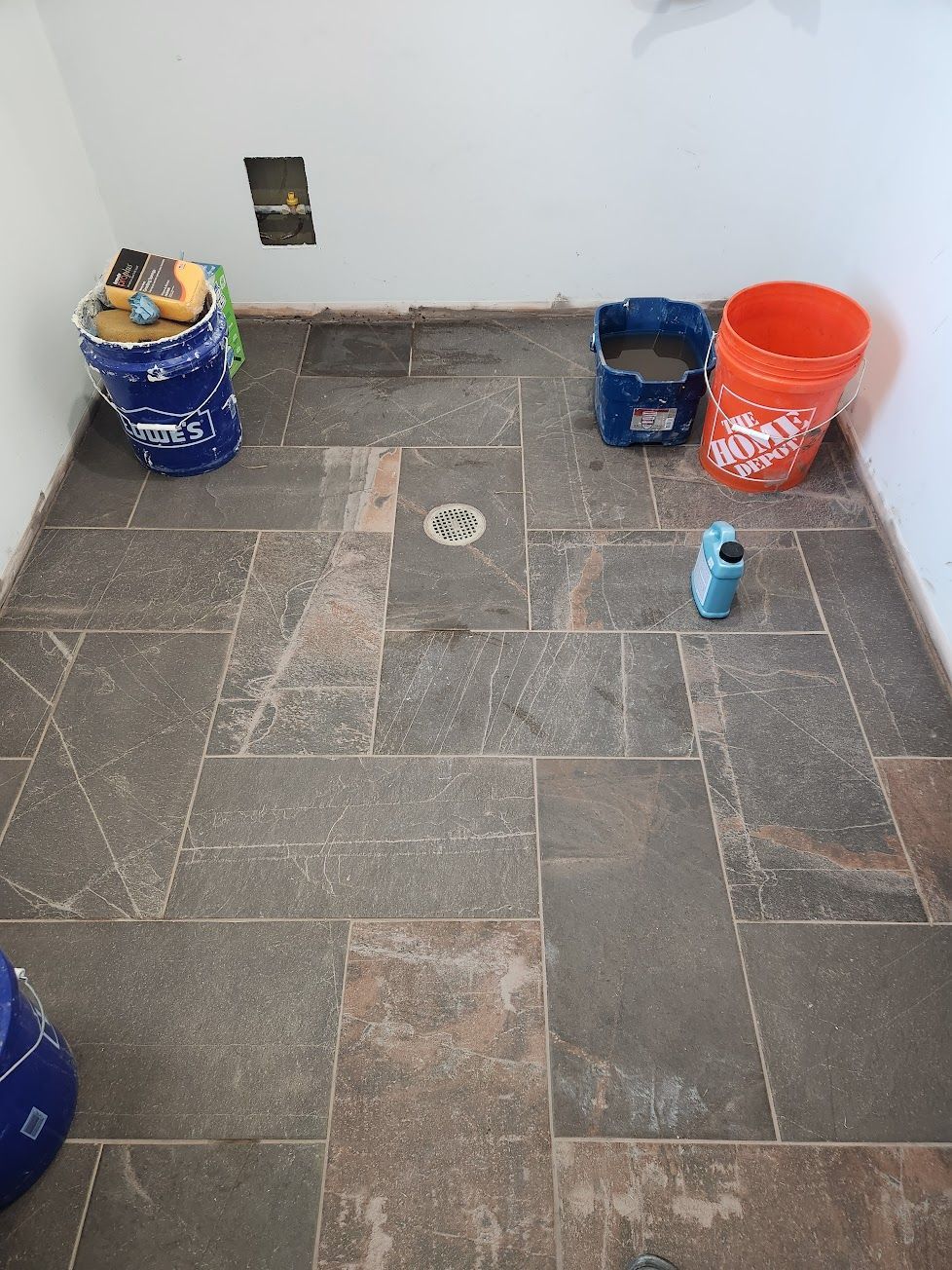 A tiled floor in a construction area with two buckets, a sponge, and a small bottle of cleaner near a center drain.