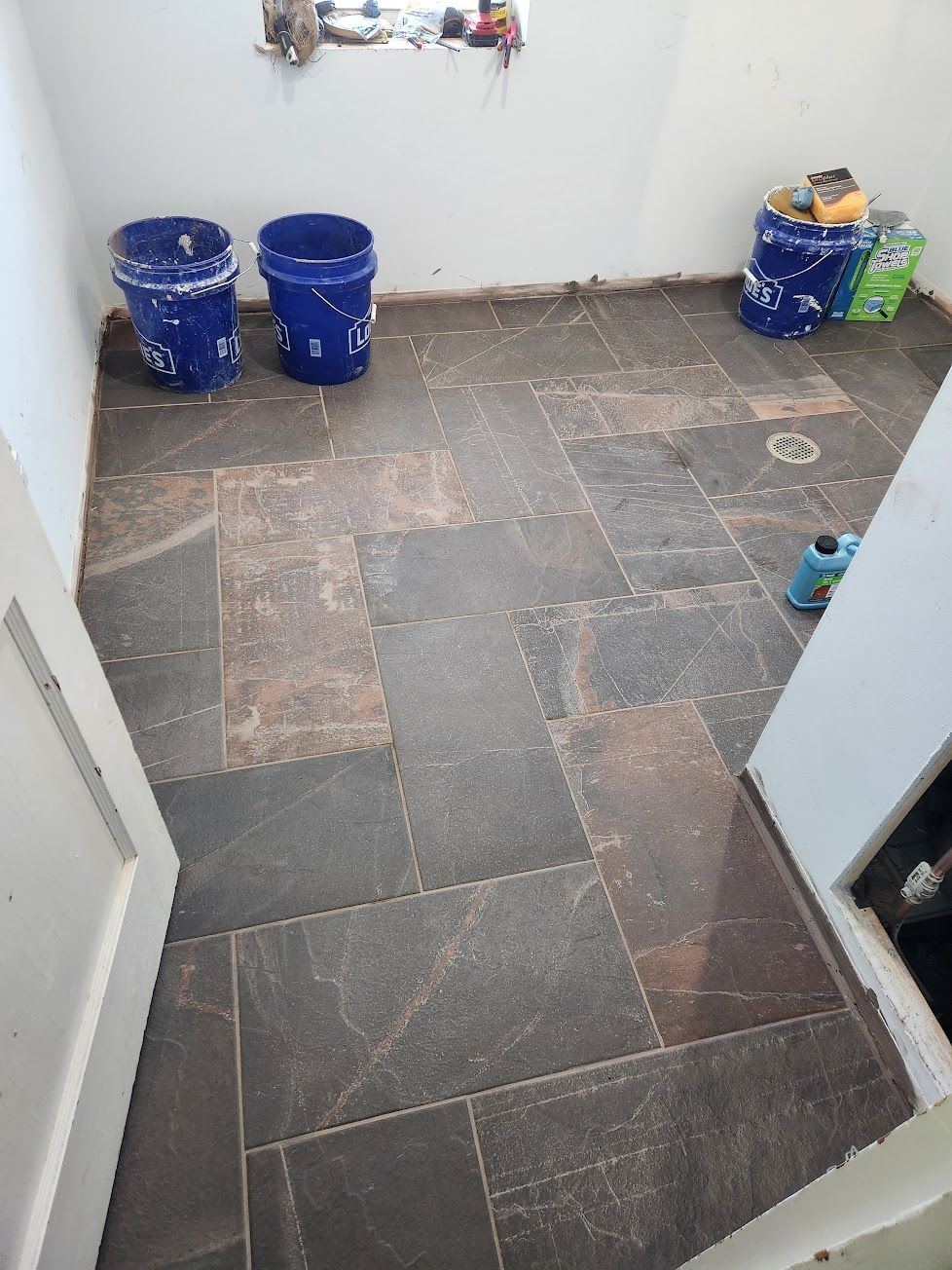 A room with dark, rectangular stone floor tiles, two blue buckets in the corner, and supplies near a door frame.